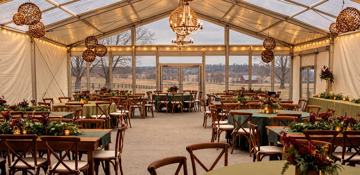 Clear-top frame tent set for a holiday event with chandeliers, farm tables and greenery